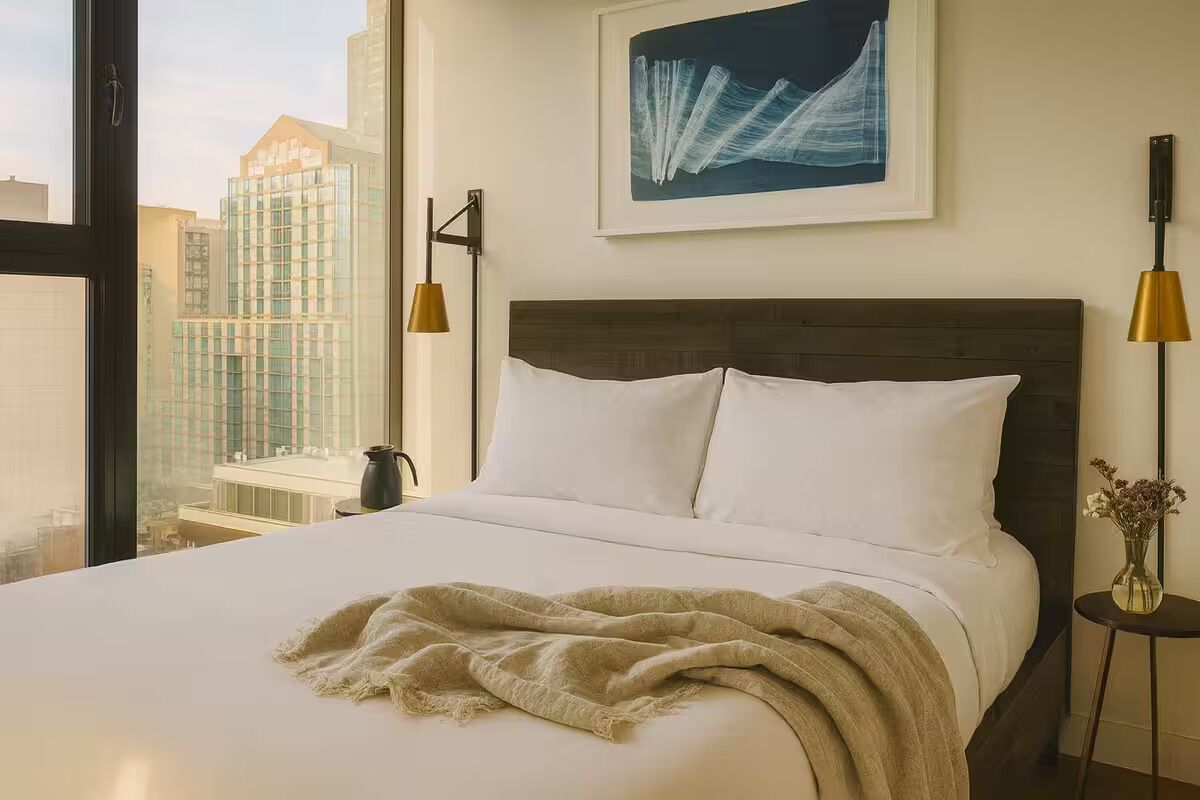 A hotel room with a large bed, a city view, a framed abstract painting, and two bedside lamps.