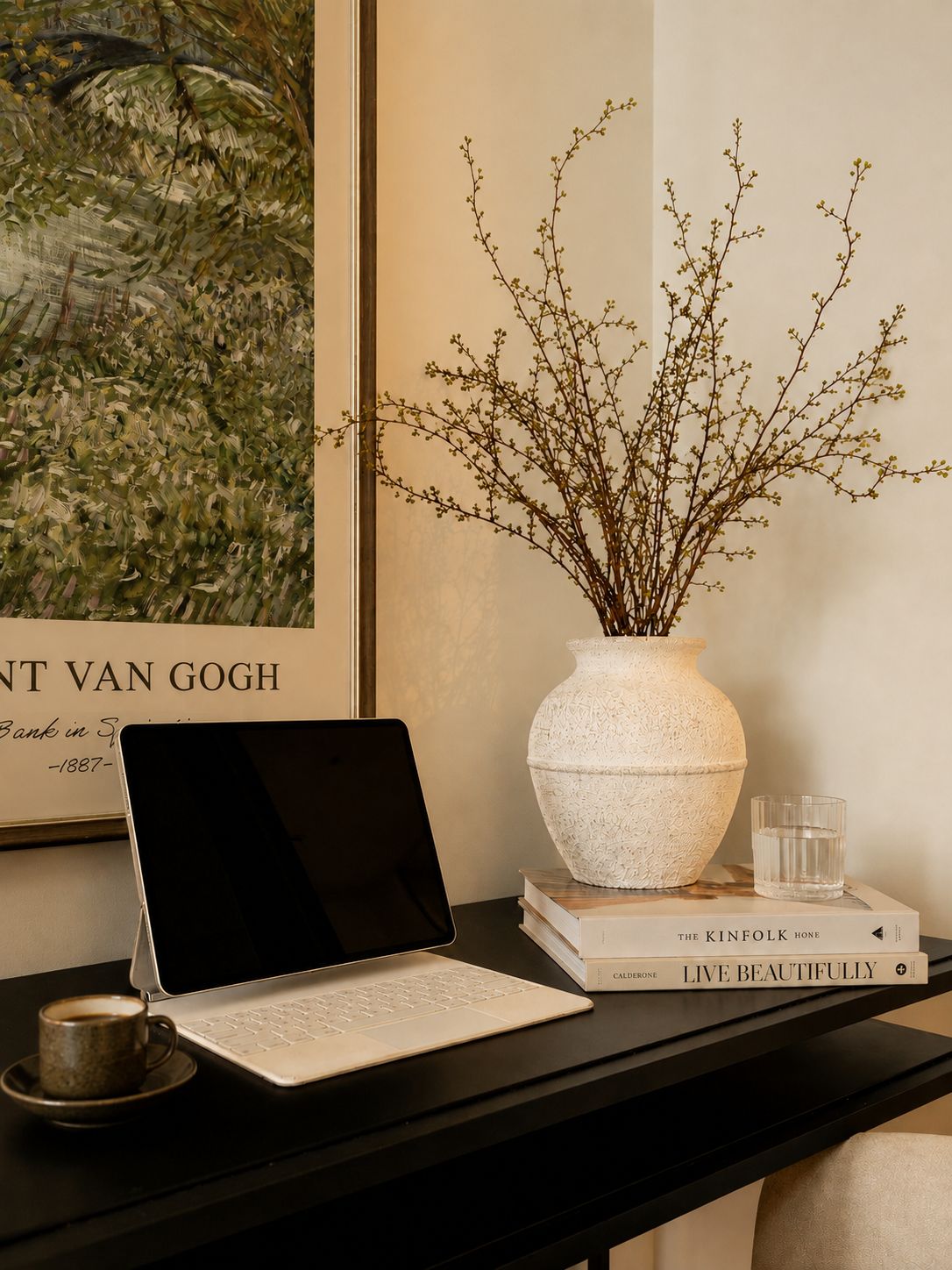 A laptop on a desk with a vase of branches, a glass of water, and books.