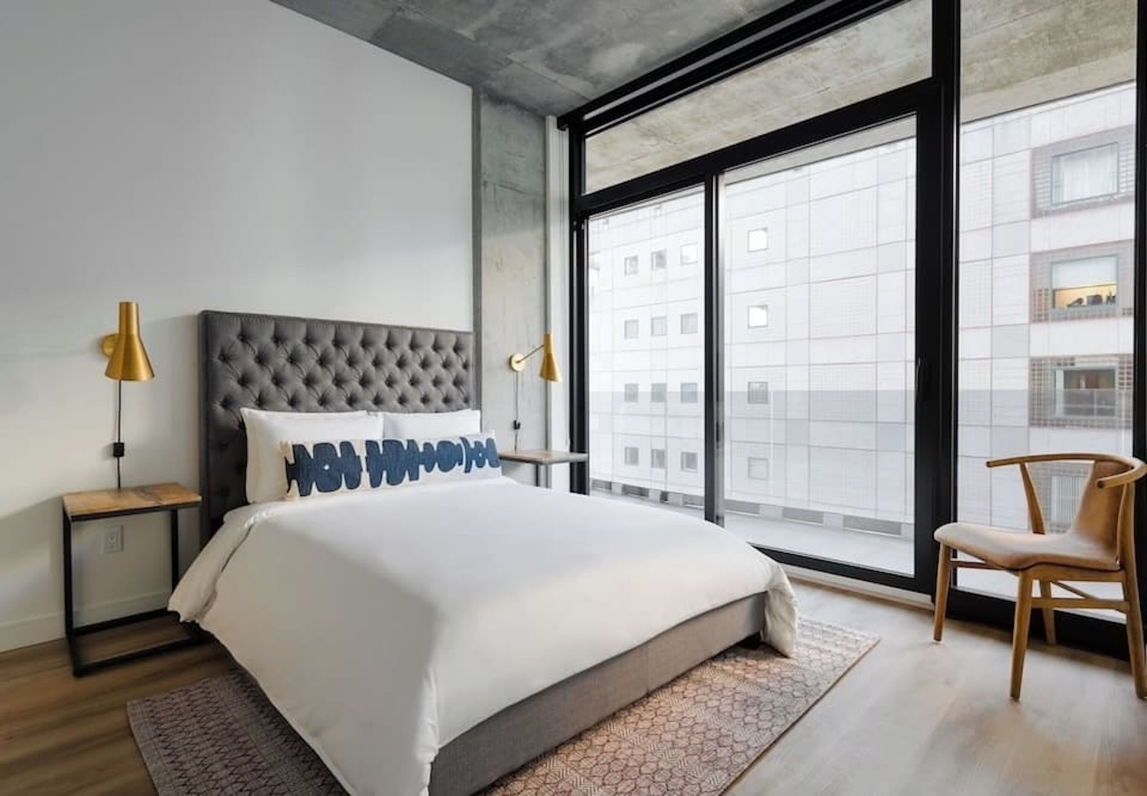 A modern bedroom with a tufted headboard, a large bed, a view of buildings through floor-to-ceiling windows, a small wooden nightstand, and a chair.