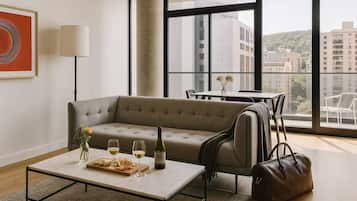 A modern living room with a sofa, a coffee table with snacks and wine, a lamp, and a view of buildings through large windows.