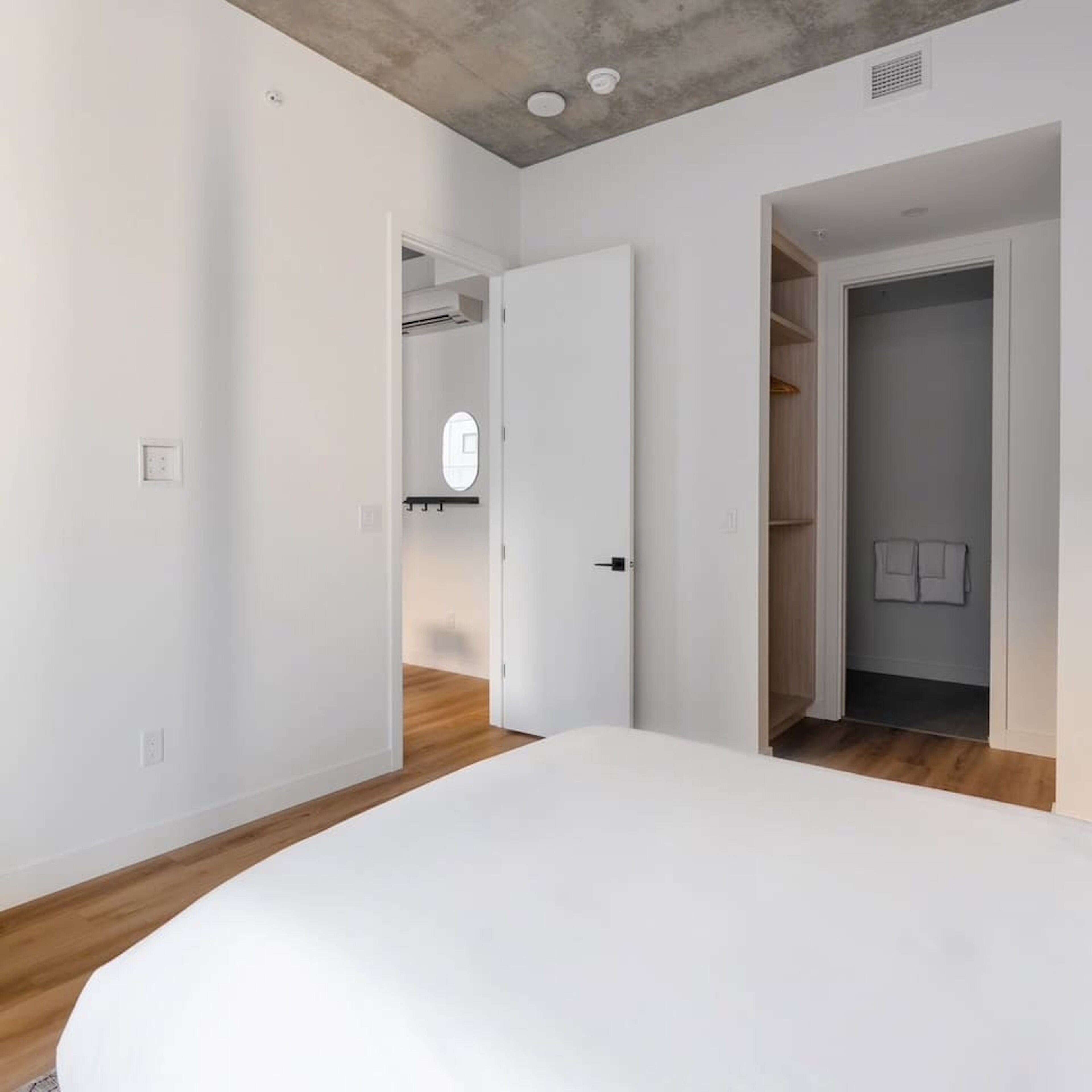 A modern bedroom with a large bed, wooden flooring, white walls, and a closet with hanging clothes.
