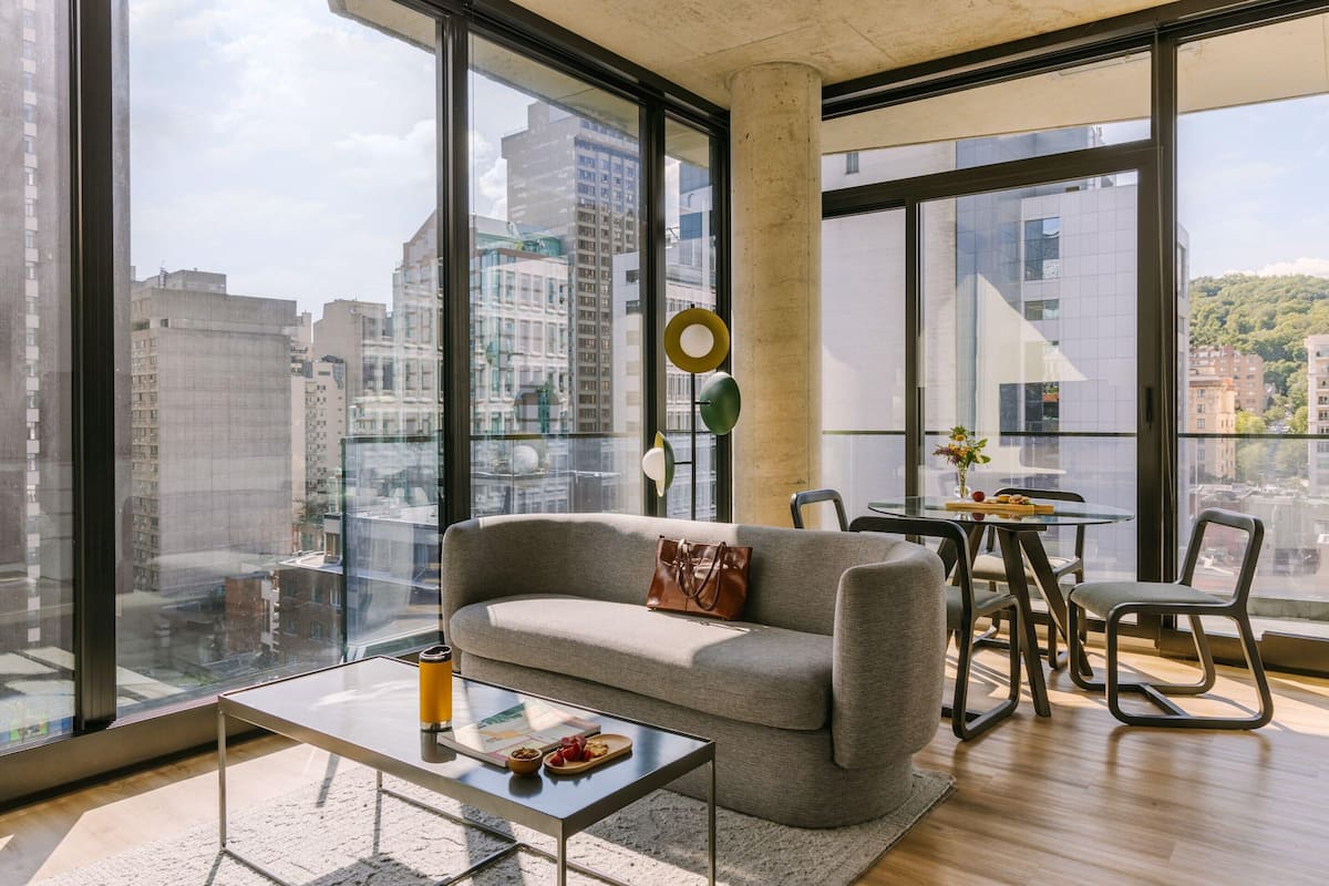 Una sala moderna con sofá, mesa de centro y comedor, ventanas amplias con vista a la ciudad y una lámpara circular.