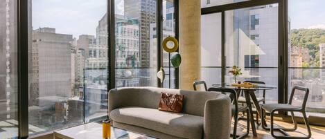 A modern living room with a sofa, coffee table, and dining area, large windows with a city view, and a circular light fixture.