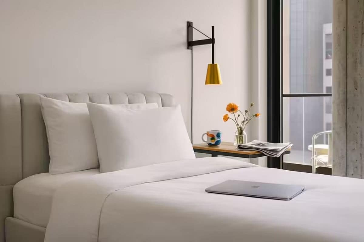 A modern hotel room with a bed, a laptop, a bedside table with a mug, and a window with a view of buildings.