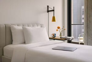 A modern hotel room with a bed, a laptop, a bedside table with a mug, and a window with a view of buildings.