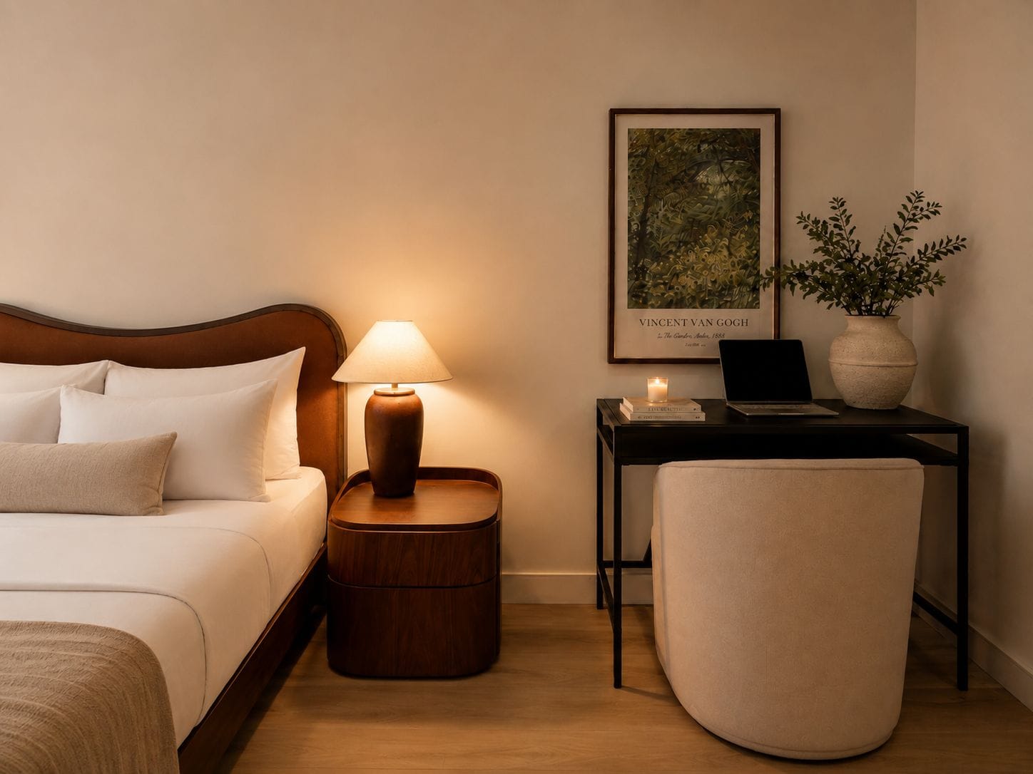 A hotel room with a bed, bedside lamp, framed artwork, a small table with a laptop, and a potted plant.