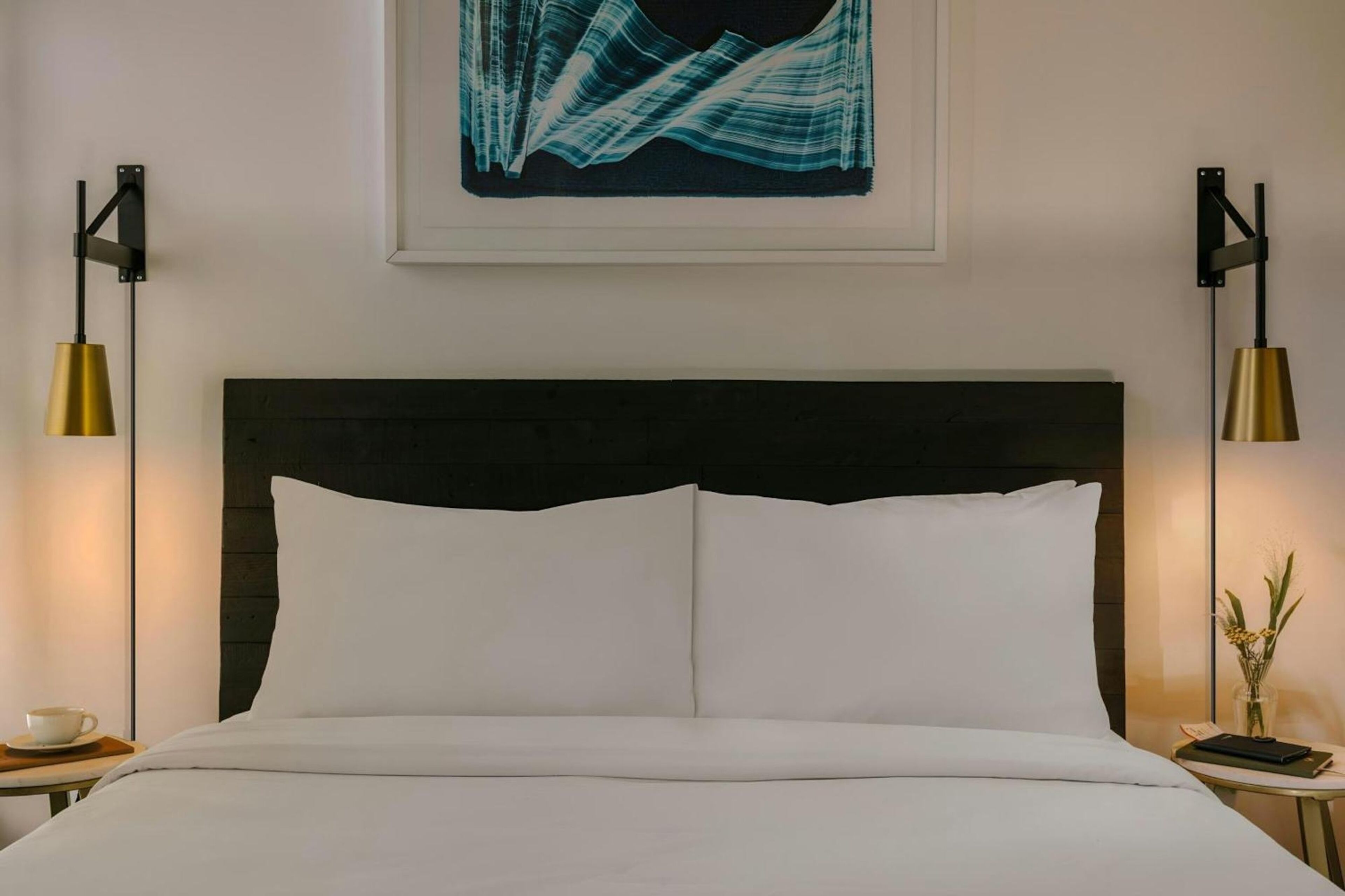 A hotel room with a bed, two bedside tables, a framed artwork above the bed, and two wall-mounted lamps.
