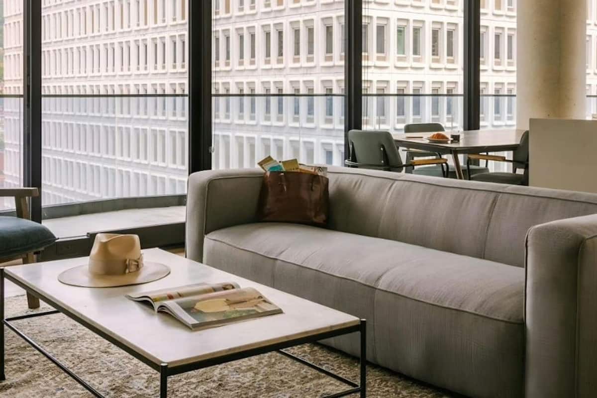 Una sala de estar moderna con un sofá, una mesa de centro y vistas a un paisaje urbano a través de amplios ventanales.