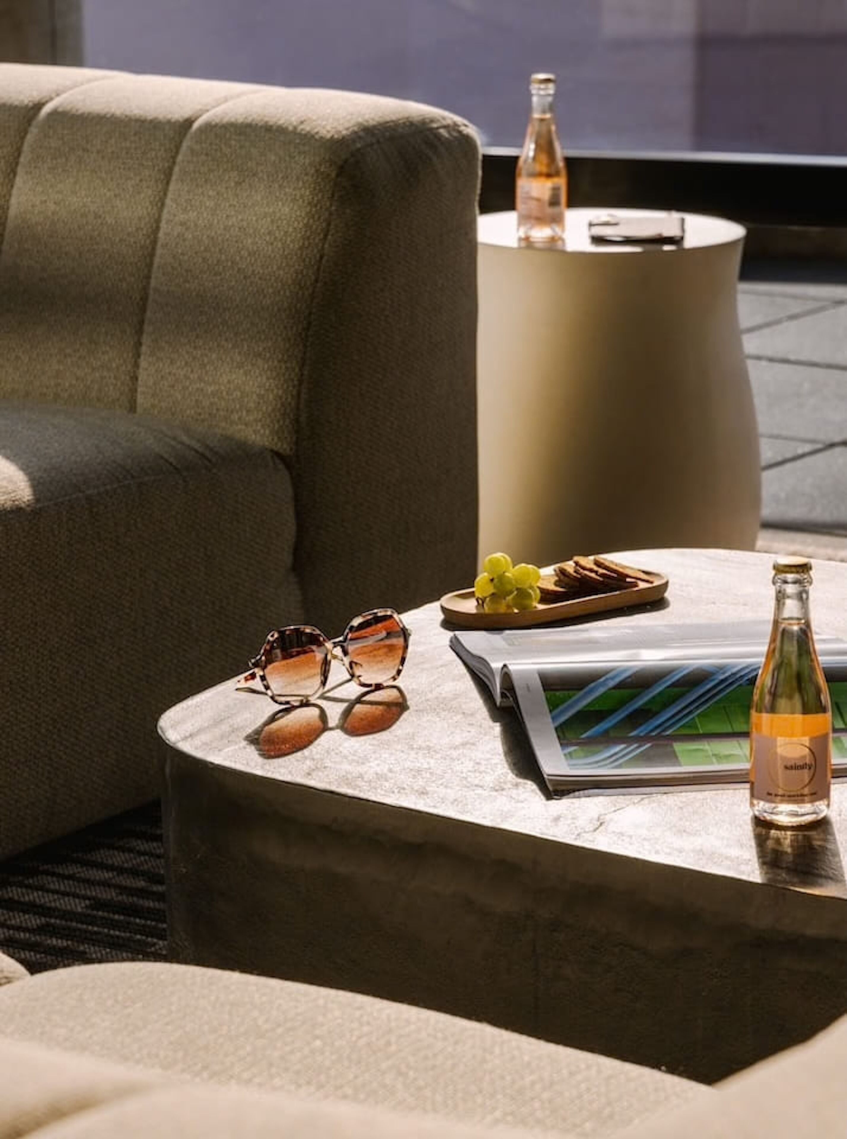 A modern lounge area with a sofa, a round table with a magazine, a bottle, and a tray with snacks.