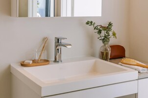 Une salle de bain avec un lavabo blanc, un robinet chromé et un miroir au-dessus. On y trouve un plateau en bois avec une brosse à dents et un verre, ainsi qu’un vase avec des plantes vertes sur le comptoir.