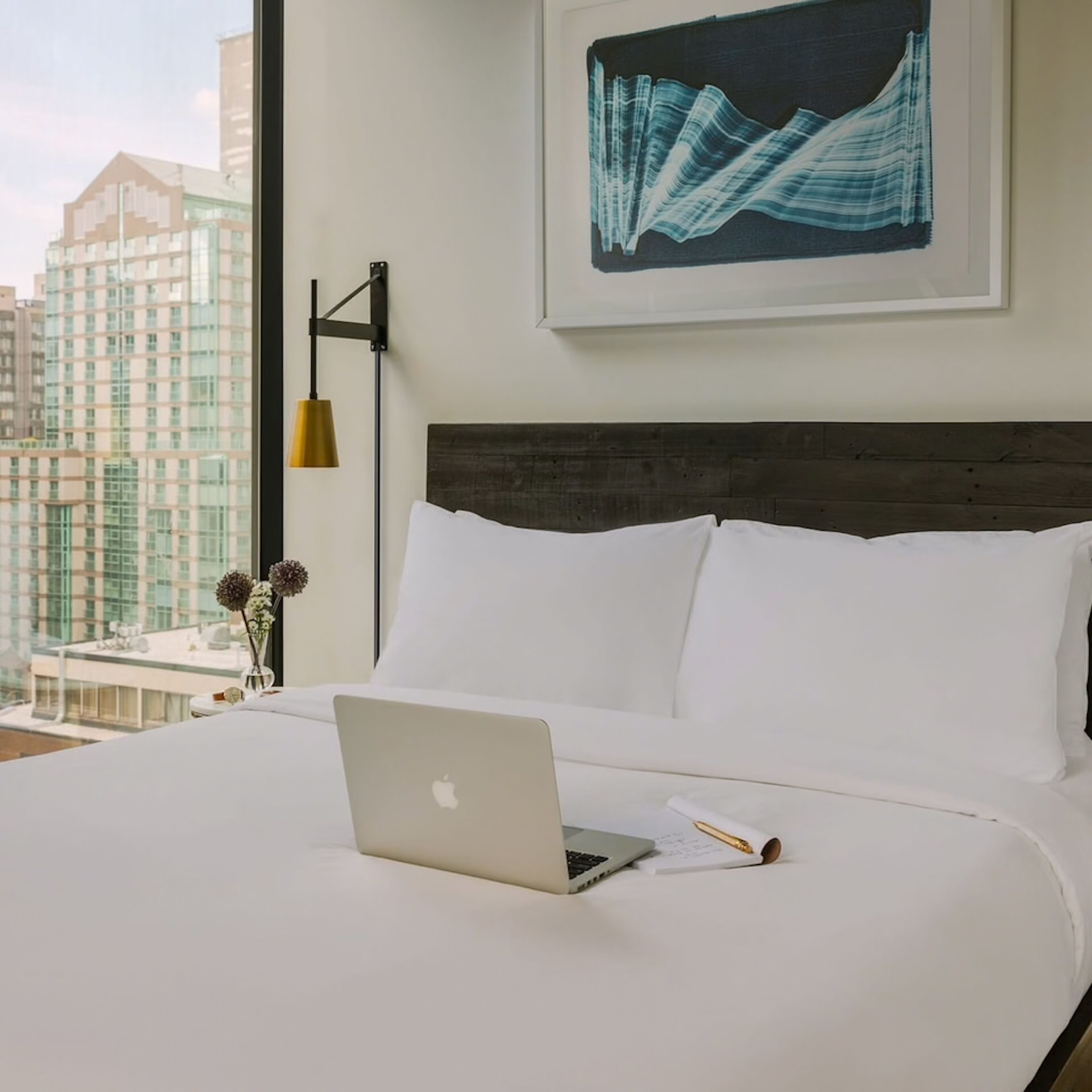 A hotel room with a bed, a laptop, a pen, and a framed artwork on the wall.