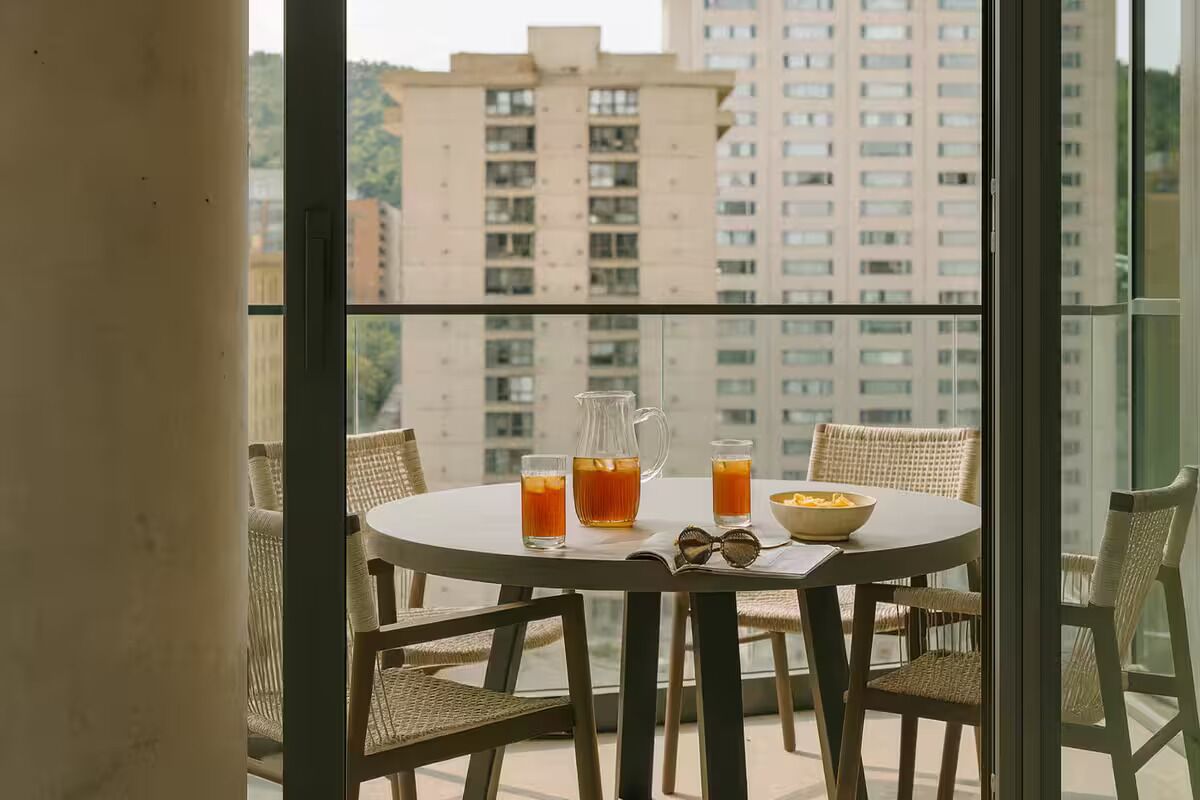 A round table with wicker chairs, a pitcher, glasses, and a bowl on a balcony with a view of high-rise buildings.
