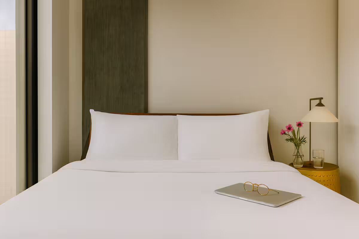 Una habitación de hotel con una cama, una mesita de noche con una lámpara, y un ordenador portátil sobre la cama.