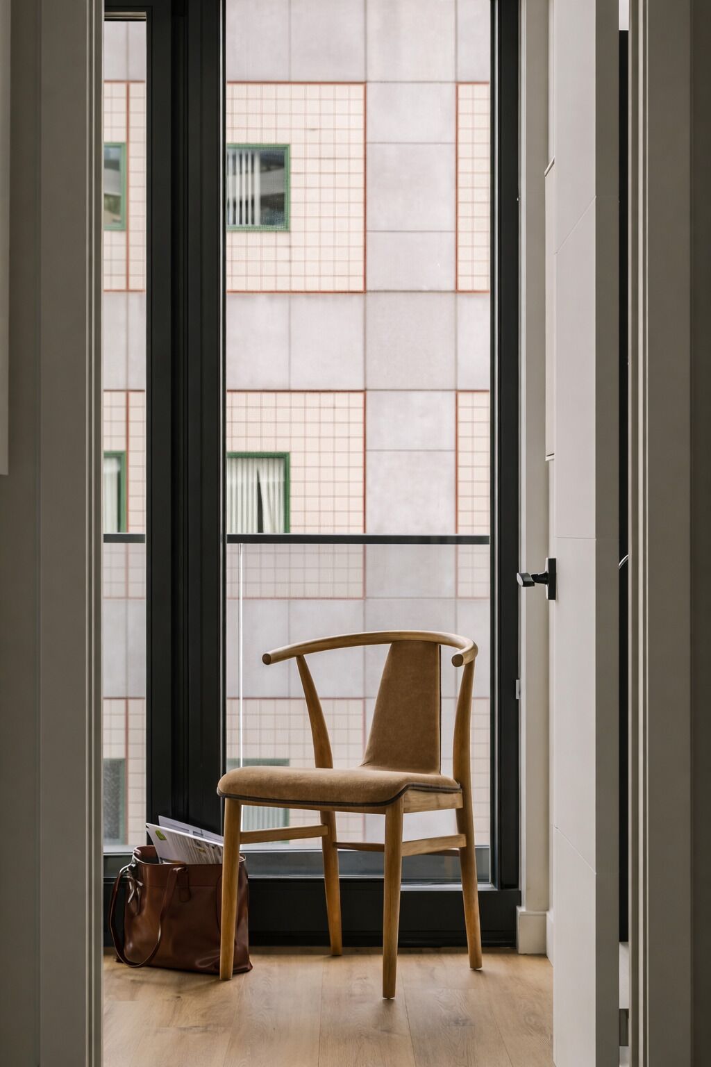 Ein Zimmer mit einem Holzstuhl, einer braunen Ledertasche und einem Fenster mit Blick auf ein Gebäude mit Gitterstruktur.