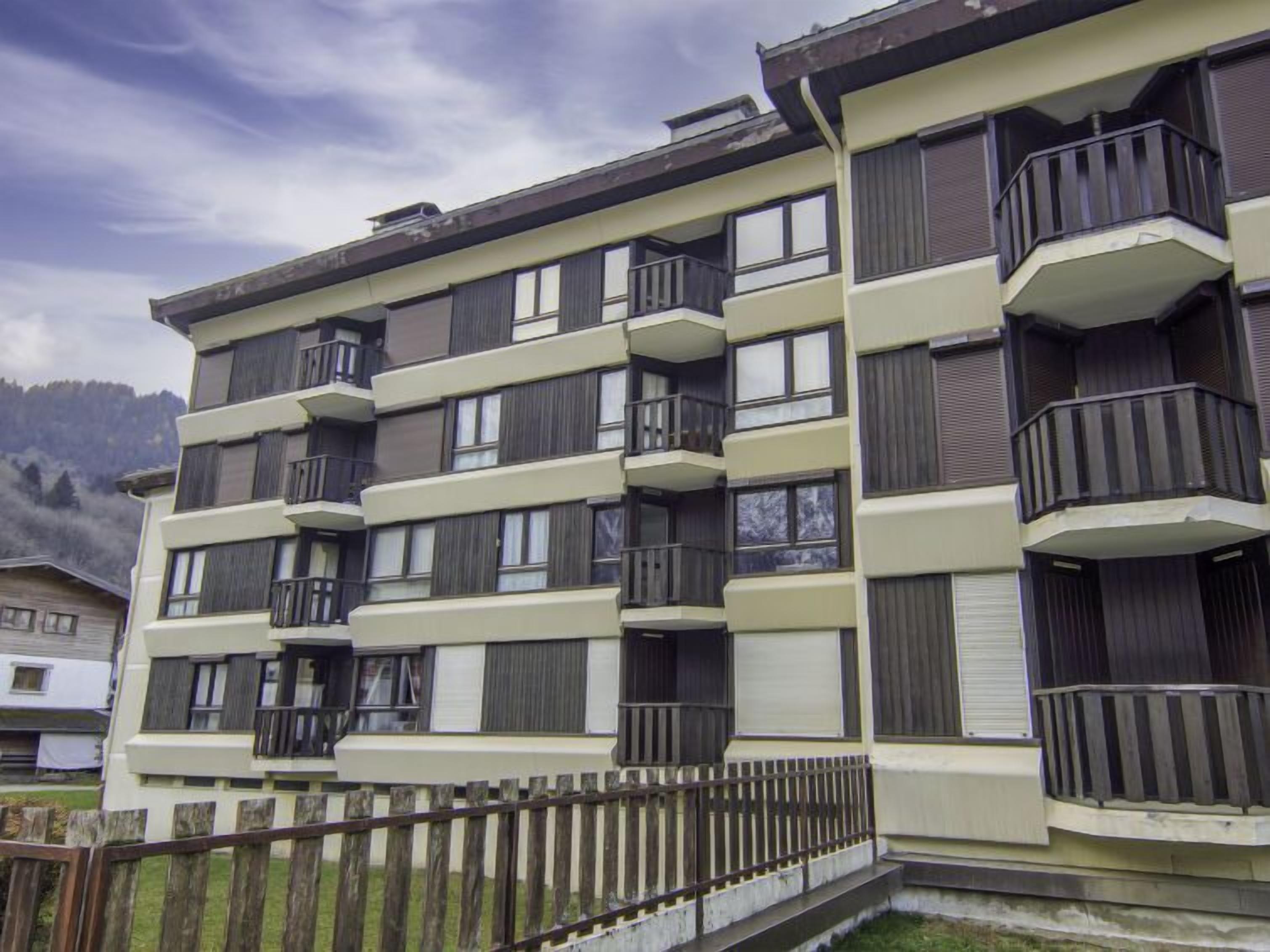 A multi-story residential building with balconies, a wooden fence, and a mountainous backdrop.
