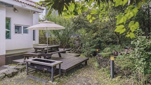 An outdoor patio area with picnic tables and an umbrella, surrounded by greenery and a stone wall. - Jeju Supgol Dokchae Pension (Jeju City)
