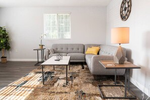 A living room with a grey sofa, a coffee table, a lamp, a rug, a potted plant, and a window with blinds.