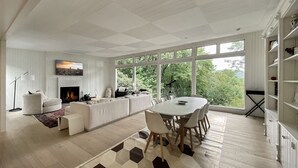 Ein modernes Wohnzimmer mit Kamin, Essbereich und großen Fenstern mit Blick auf Grünflächen.