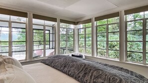 Ein Schlafzimmer mit großen Fenstern, einem Bett und Blick auf Grün.