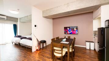 Een moderne hotelkamer met een houten vloer, een eettafel met stoelen, een aan de muur bevestigde televisie, een bed met hoofdeinde en een keukenhoek met koelkast en magnetron.