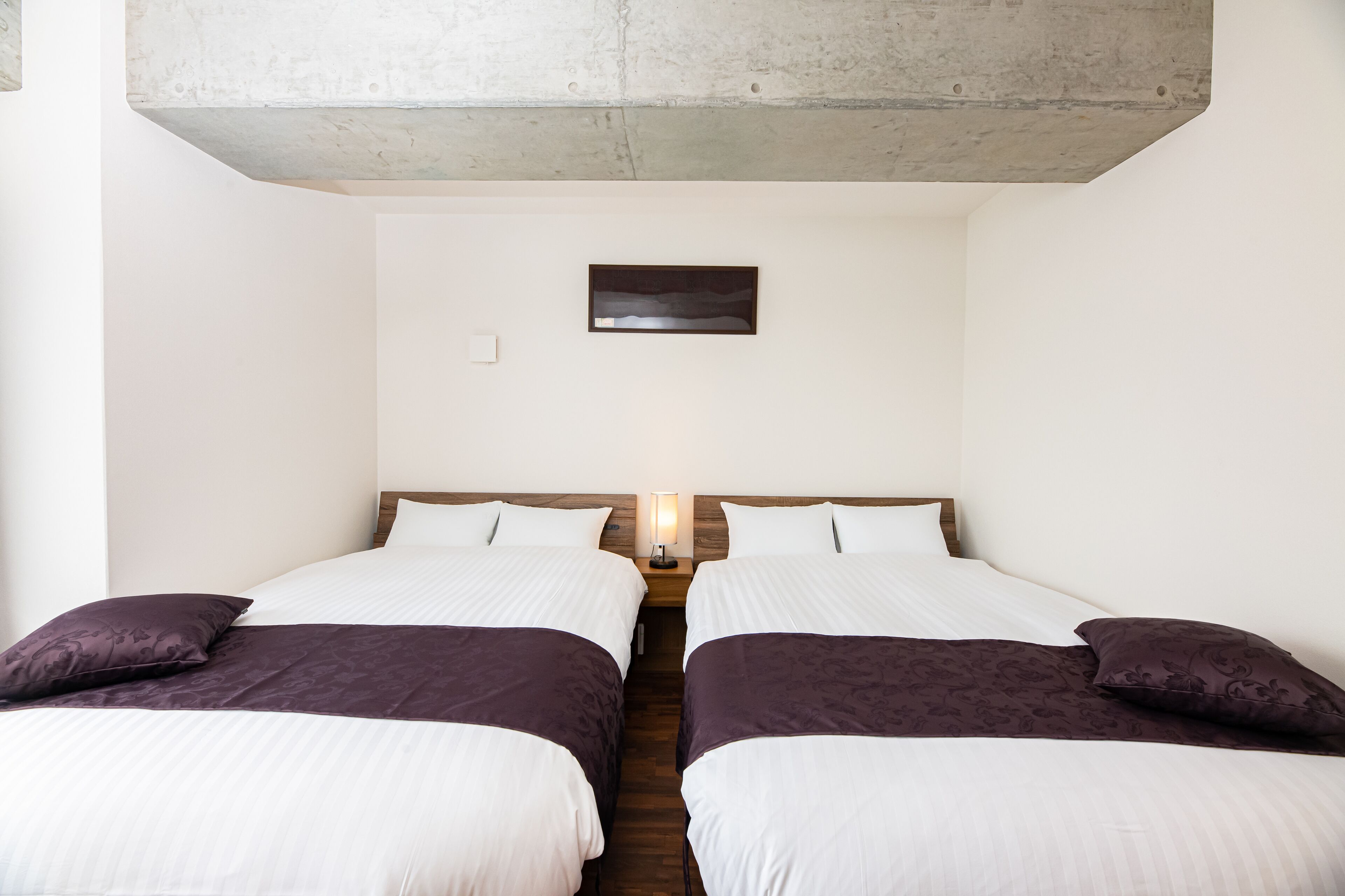 Una habitación con dos camas, una mesita de noche con lámpara, un cuadro pequeño en la pared y techo de concreto.