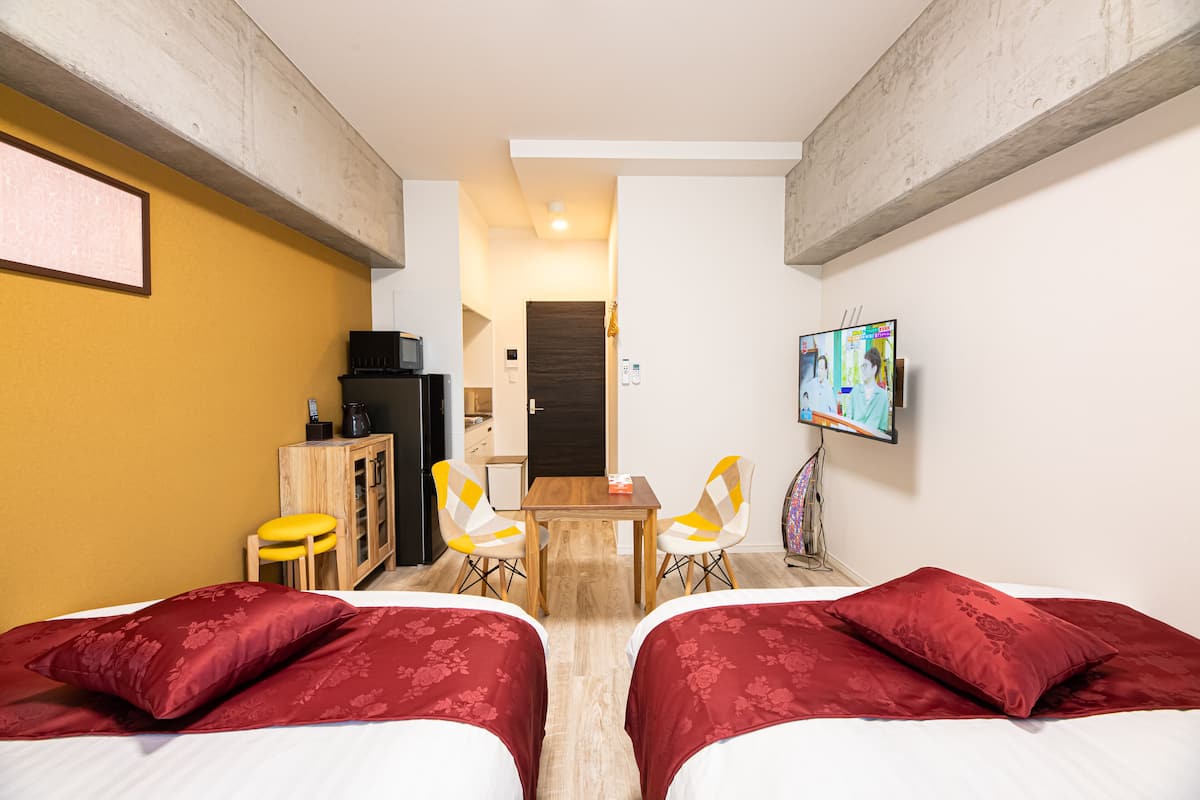 Habitación de hotel con dos camas, un escritorio, sillas, televisor y una pequeña kitchenette.