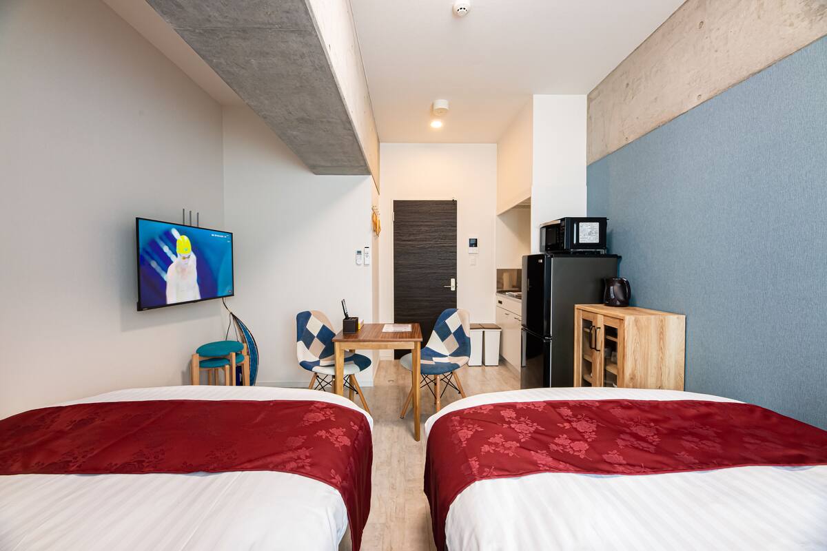 Habitación de hotel con dos camas, televisión, escritorio con sillas, kitchenette y armario.
