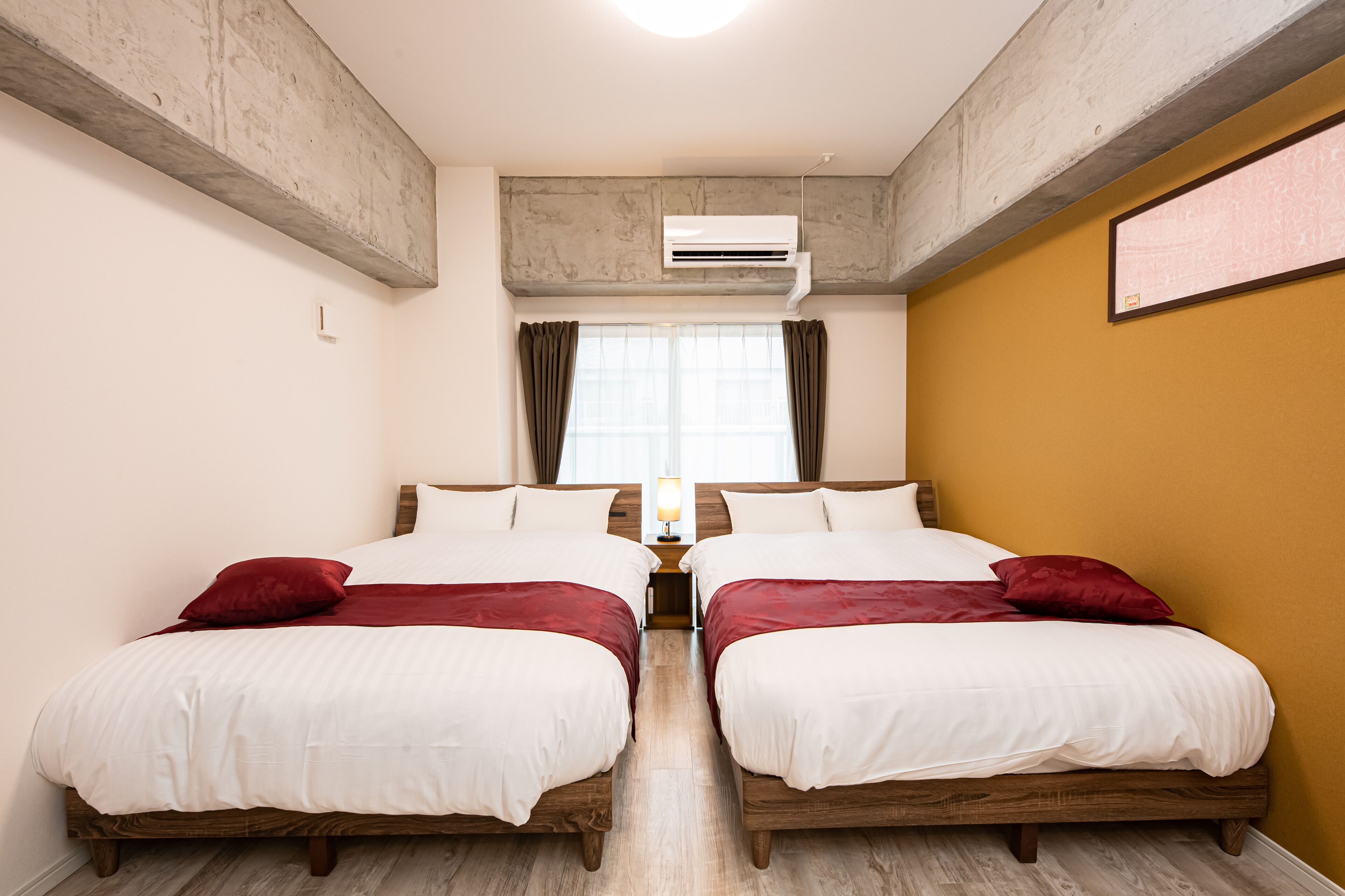 Habitación doble con dos camas, ventana con cortinas, equipo de aire acondicionado y piso de madera.