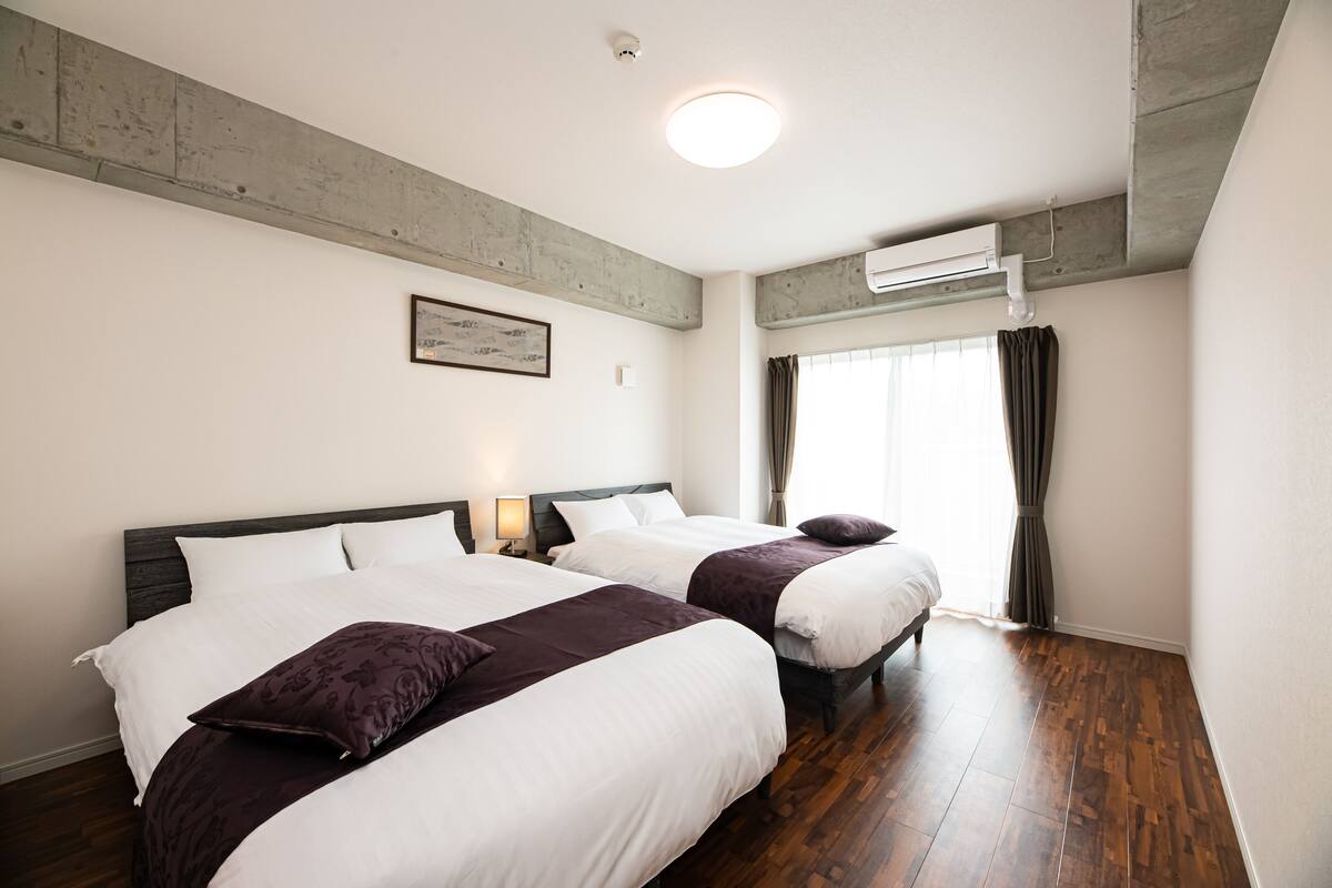 Habitación de hotel con dos camas, suelo de madera, ventilador de techo y ventana con cortinas.