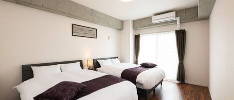 A hotel room with two beds, wooden flooring, a ceiling fan, and a window with curtains.