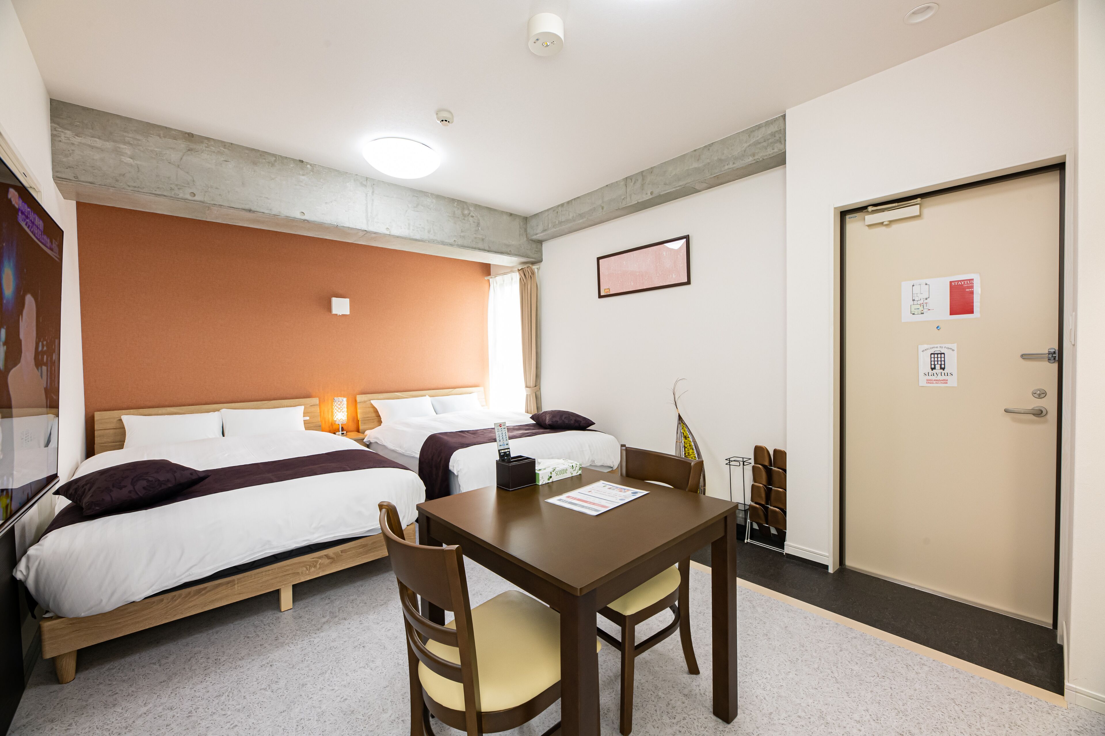A hotel room with two beds, a wooden table with chairs, a desk, and a door with a sign.