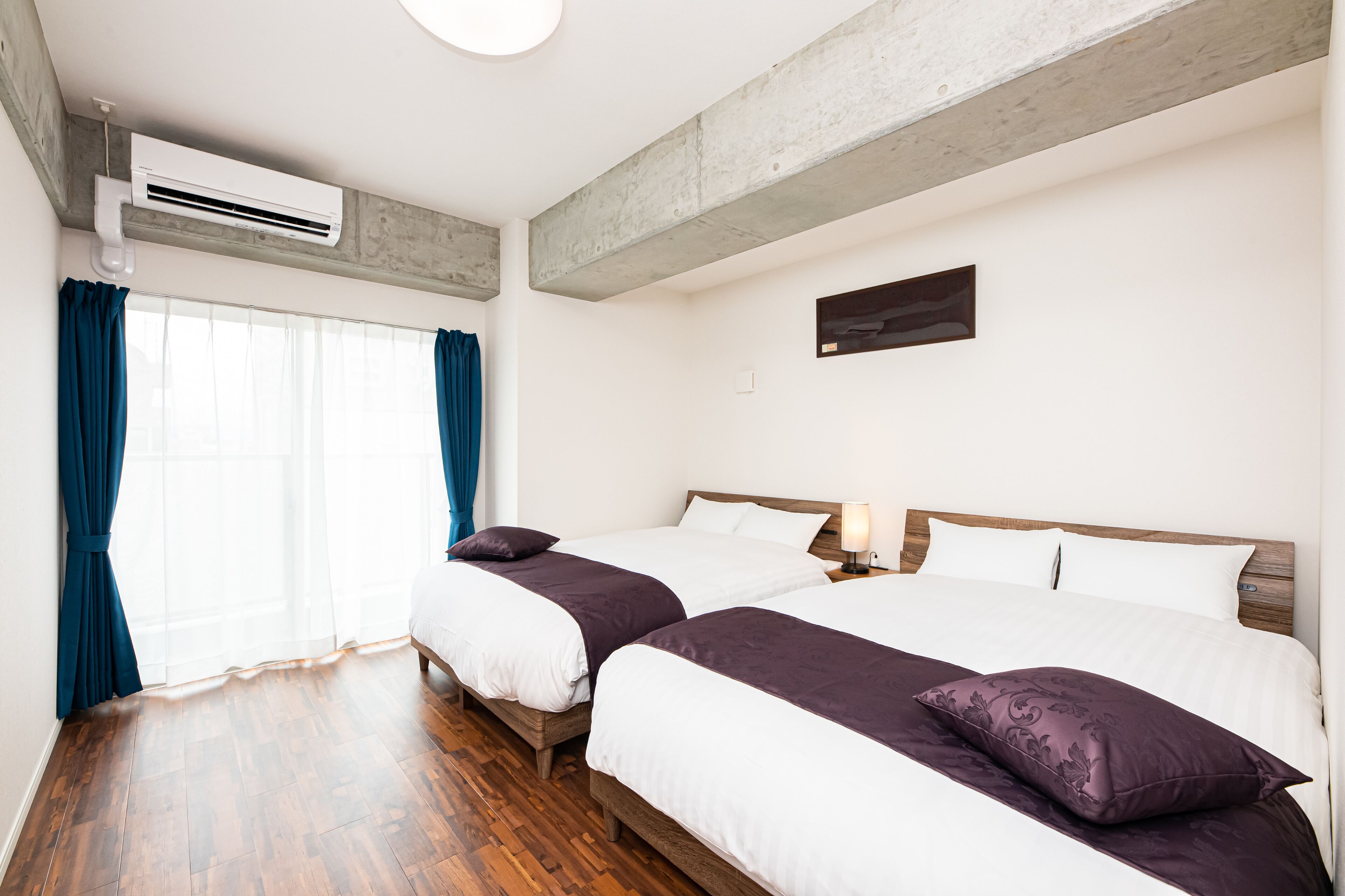 A hotel room with two beds, wooden flooring, a large window with curtains, an air conditioning unit, and a small wall-mounted picture.