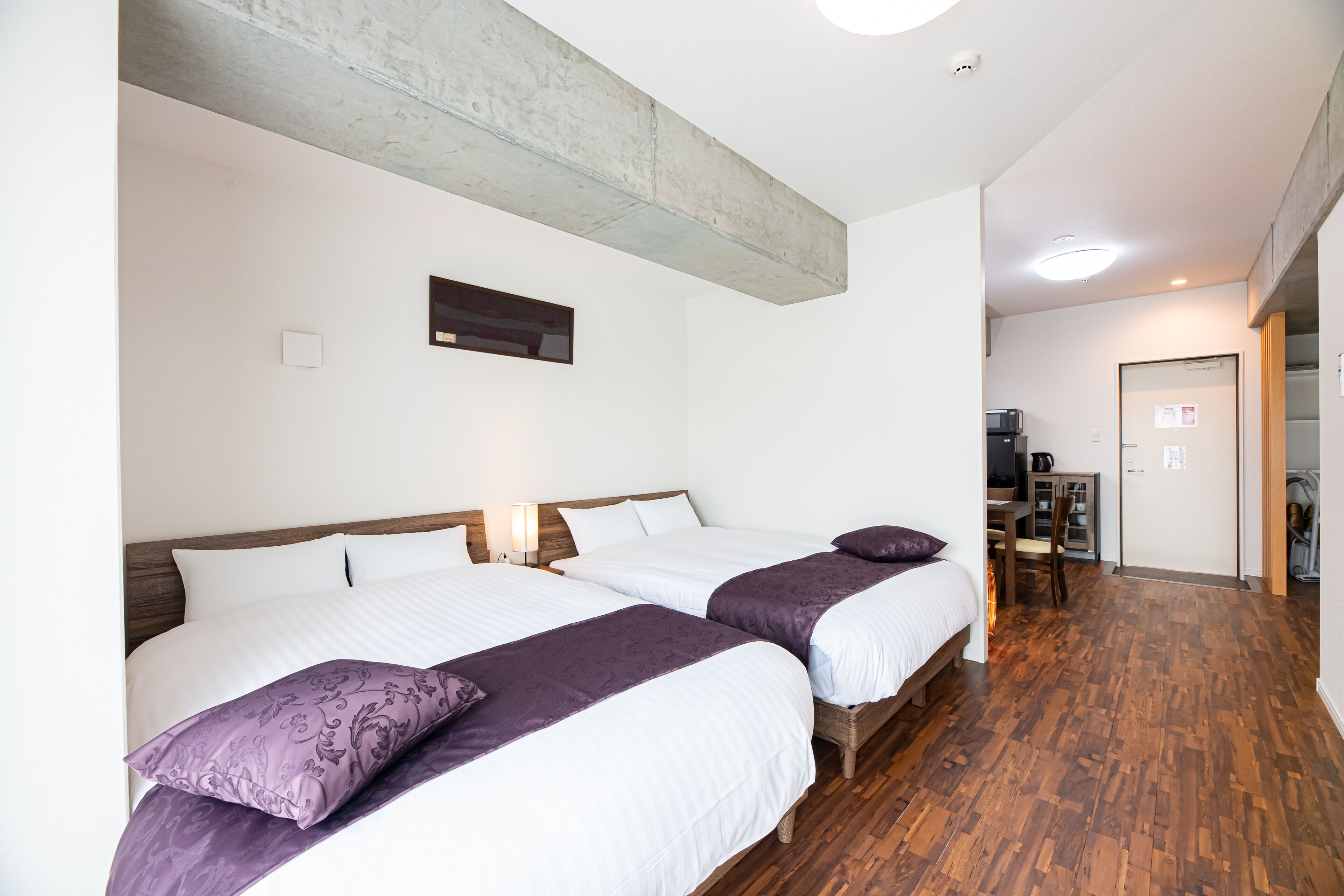 A hotel room with two beds, wooden flooring, a visible kitchen area, and a door leading to another room.