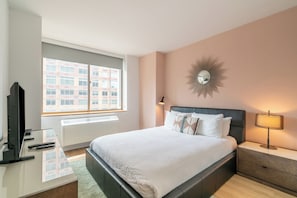 A modern hotel room with a large bed, bedside table, lamp, and a television on a glass desk.