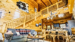 A wooden cabin interior with a loft bedroom, a dining area, and a living room with a sofa and coffee table. - Family-Friendly• Stunning Views • 7 Mi to Dollywood •Hot Tub• Highly Rated! (Sevierville)