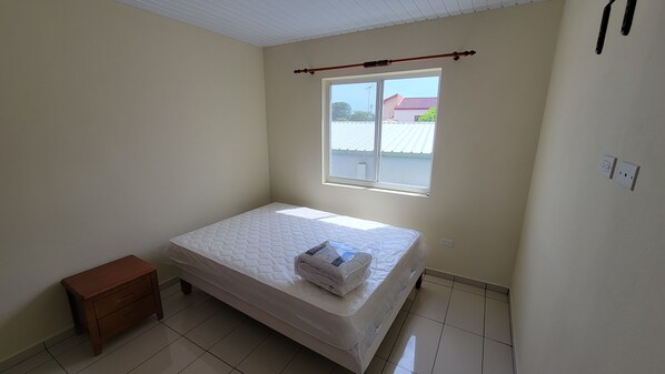 Une petite chambre propre, avec un lit simple, une petite table de chevet en bois, une fenêtre donnant sur un immeuble et un ventilateur de plafond.