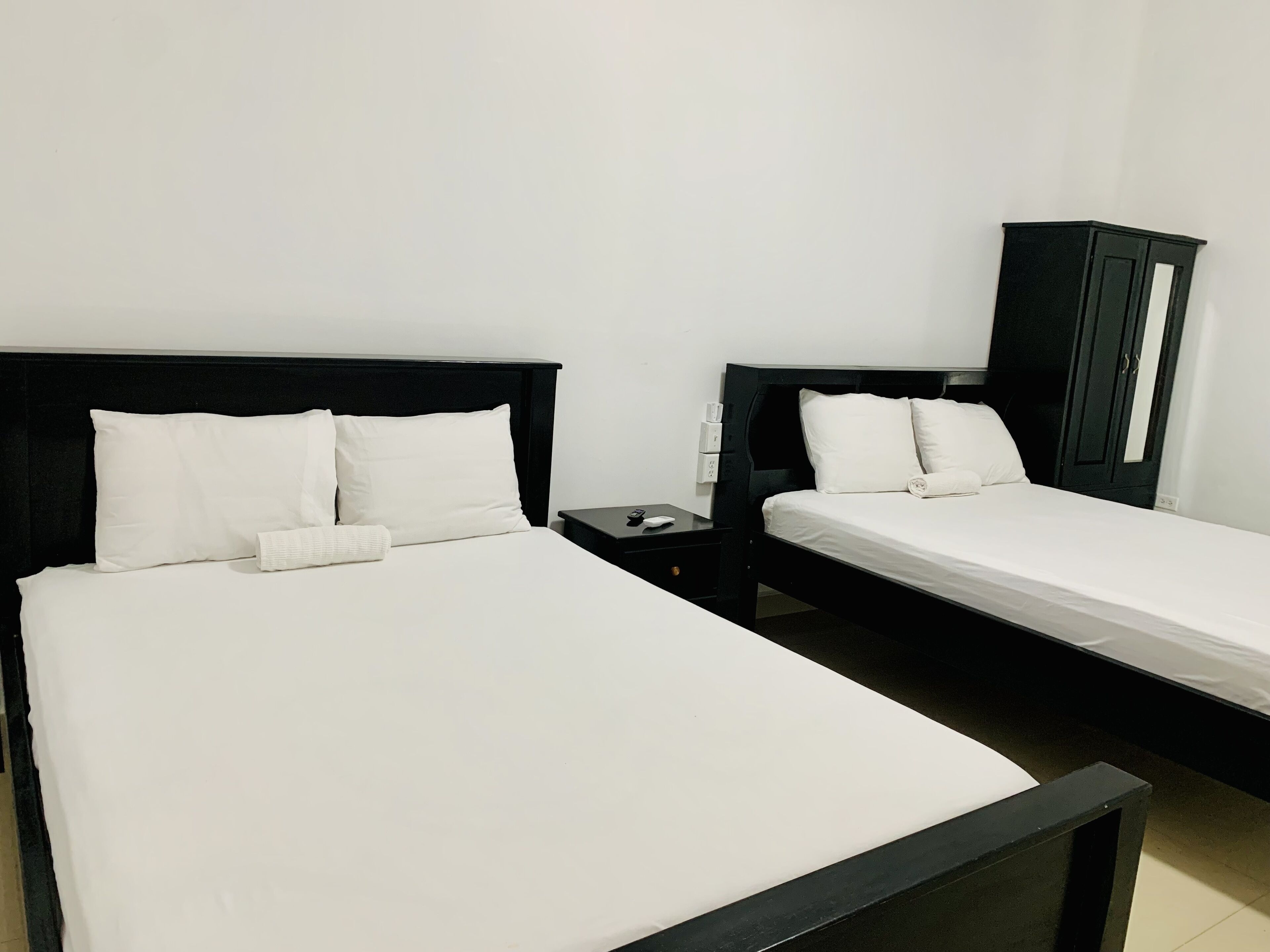 A hotel room with two single beds, each with white linens and black headboards. There is a small bedside table with a remote control and a power outlet. A black wardrobe is visible in the background.