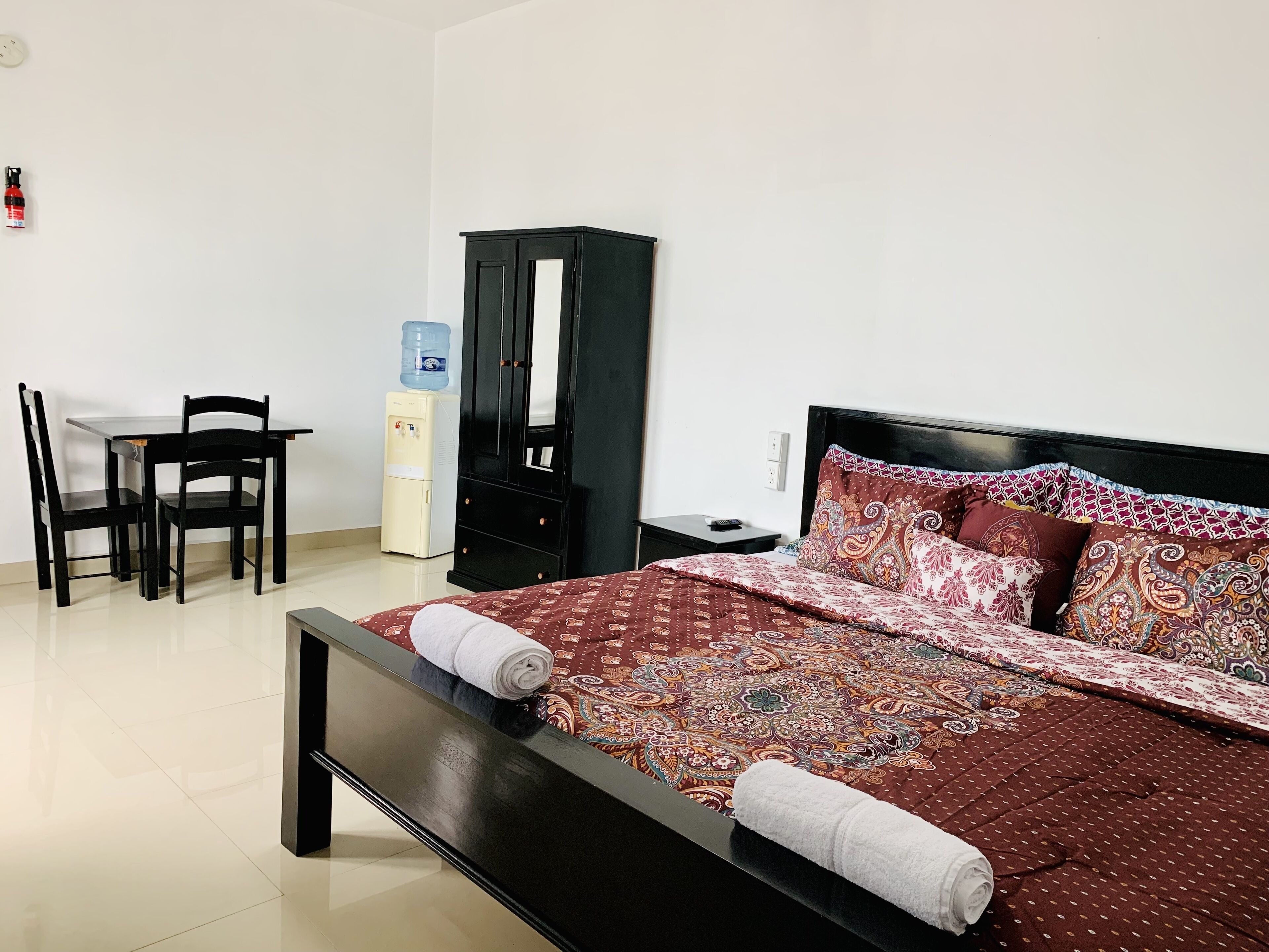 A hotel room with a bed, a desk with chairs, a water dispenser, and a wardrobe.