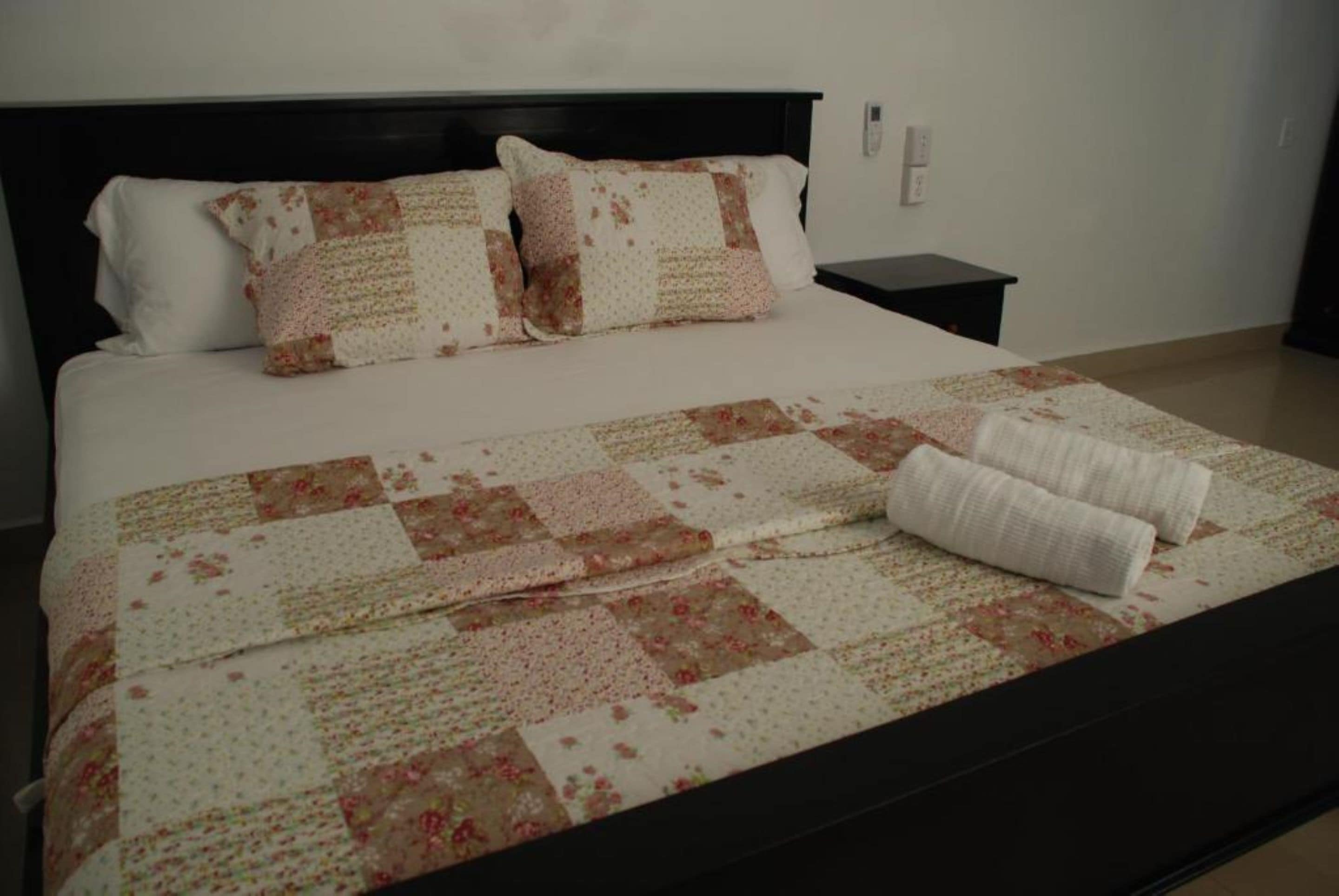 Una cama bien hecha con colcha floral y dos almohadas, una mesita de noche y un enchufe de pared.