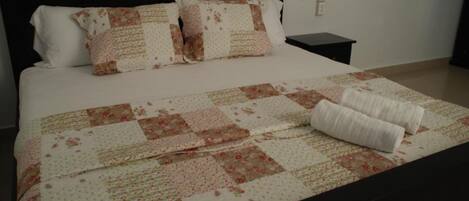 A neatly made bed with a floral quilt and two pillows, a bedside table, and a wall socket.
