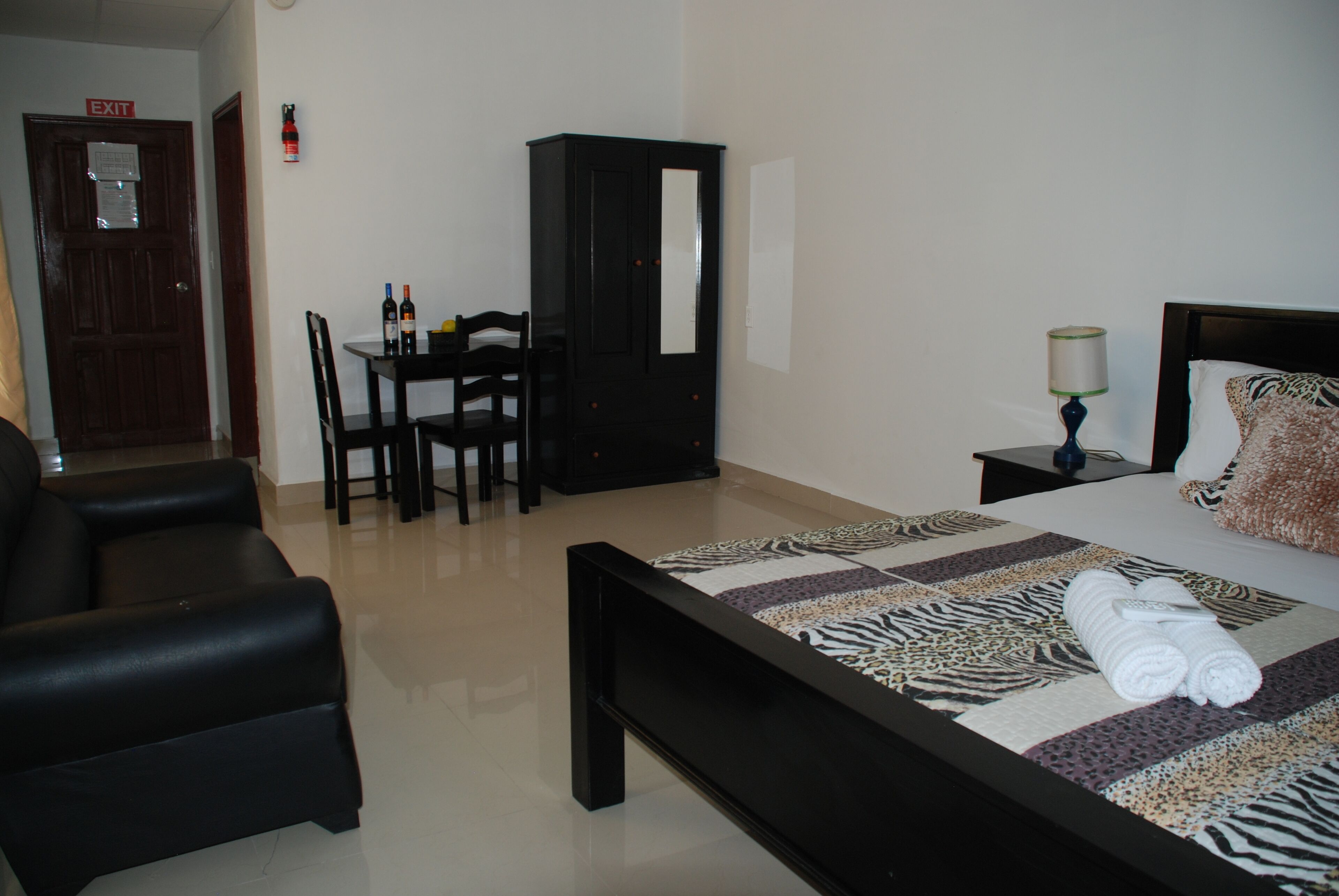 Una habitación de hotel con una cama, un escritorio con sillas, un armario y una mesita de noche con lámpara.