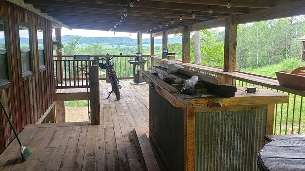 A covered porch with a wooden deck, a barbecue grill, and a view of a forested landscape. - Rustic Cabin on 7 Acres (Sparta)