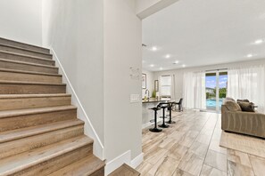 A spacious living area with a wooden staircase, a kitchen area with bar stools, and a view of a swimming pool. - Magical Memories Villa: Pop Culture, Pool, Near Disney (Kissimmee)