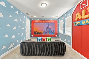 Une pièce dotée d’une grande fenêtre, d’un écran mural affichant l’image d’un château, et d’un mur rouge sur lequel on peut lire l’inscription « AL TOY BAR ».