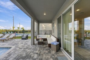 A covered outdoor seating area with a pool, lounge chairs, and a glass door leading to a poolside area. - Magical Memories Villa: Pop Culture, Pool, Near Disney (Kissimmee)