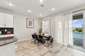 A modern kitchen with a dining area, a large window with curtains, and a view of a swimming pool.