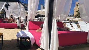 Una cama frente al mar con dosel blanco, una colcha roja y una pequeña mesa con toallas.
