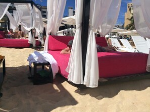 A beachside bed with white canopies, a red bedspread, and a small table with towels.