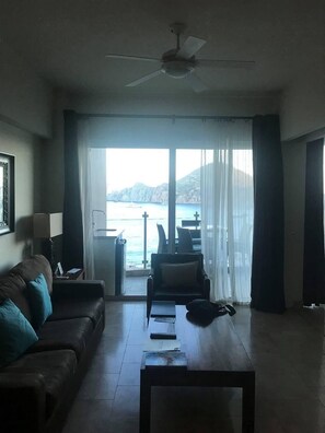 A living room with a sofa, a coffee table, and a chair. There is a large window with curtains, a ceiling fan, and a view of the sea and mountains.