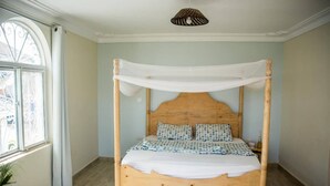 Un lit à baldaquin avec des poteaux aux quatre coins, une tête de lit en bois et une literie à motifs, dans une pièce aux murs bleu clair et dotée d’une grande fenêtre.